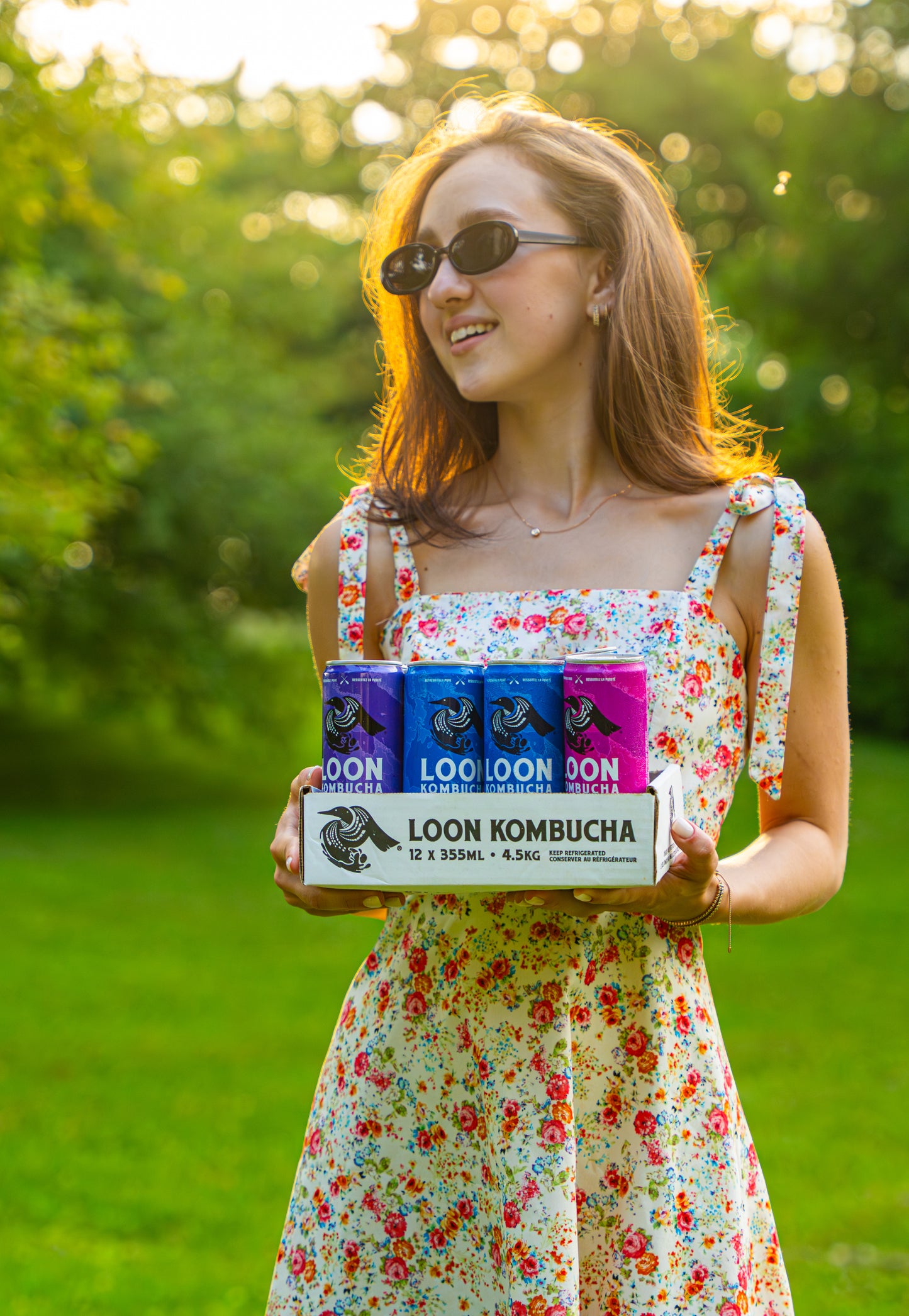 Woman holding a box of Loon Kombucha in a park setting