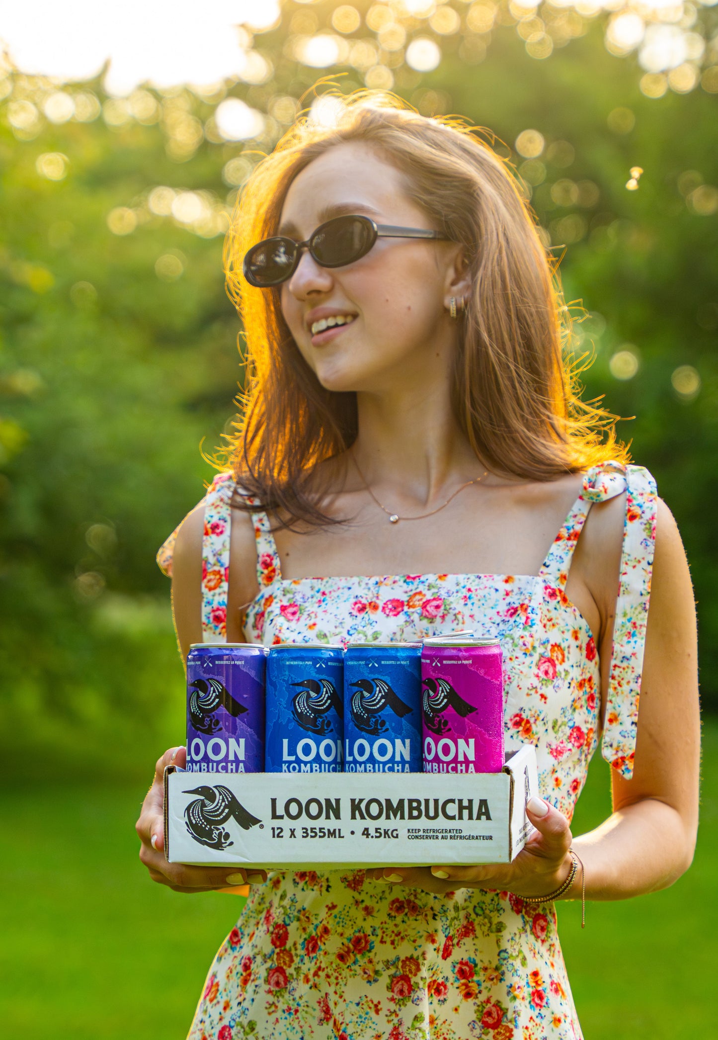 Woman holding a box of Loon Kombucha in a park setting
