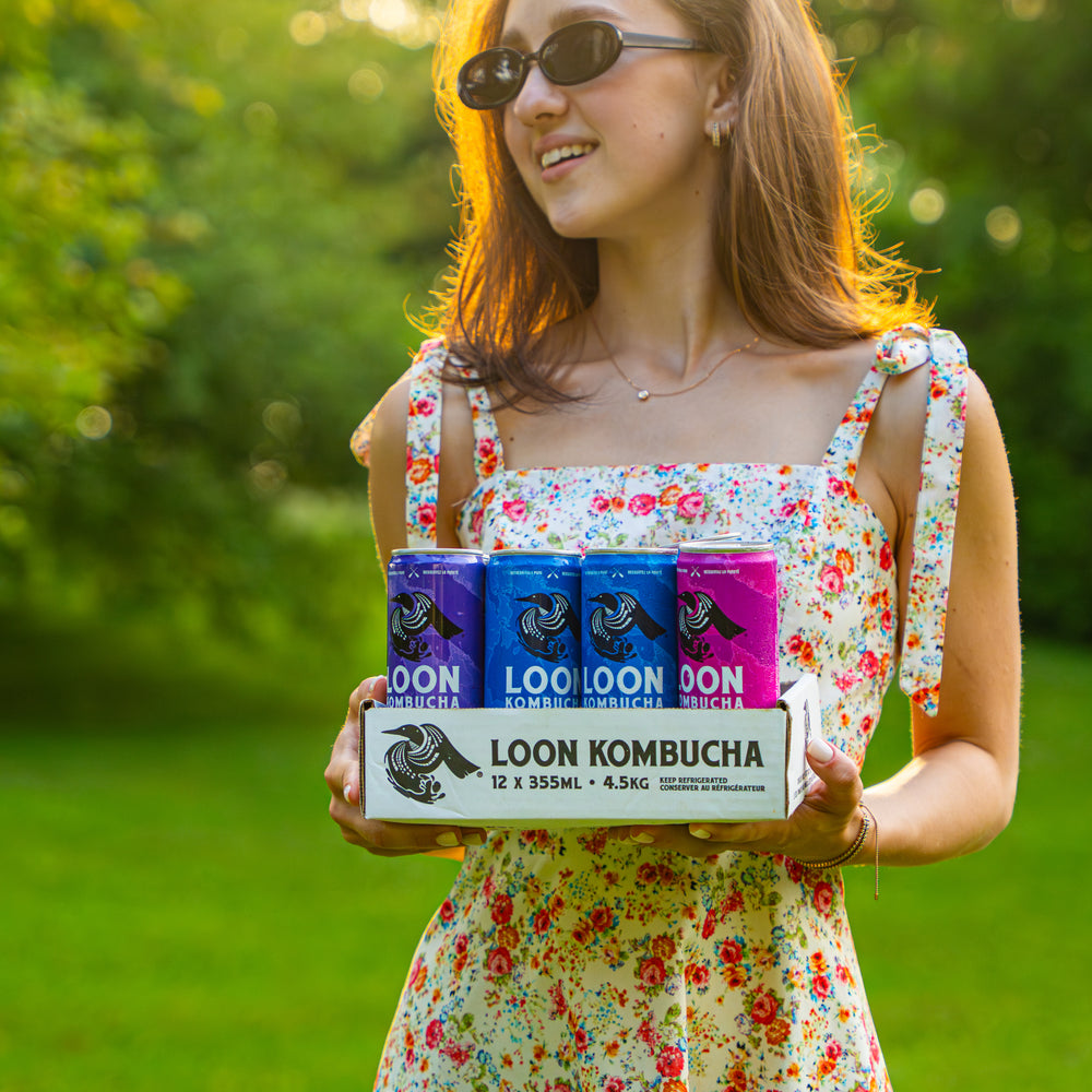 Woman holding a box of Loon Kombucha in a park setting