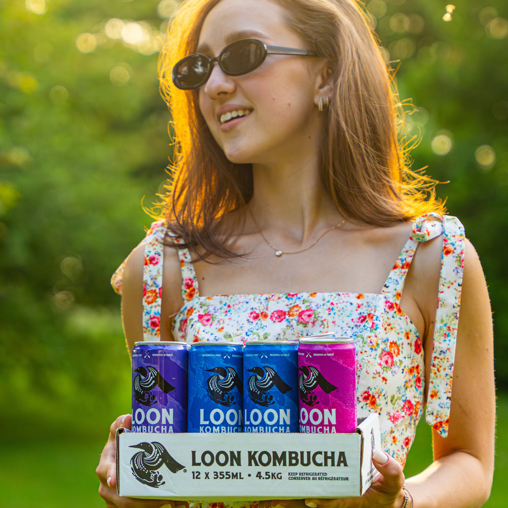 Woman holding a box of Loon Kombucha in a park setting